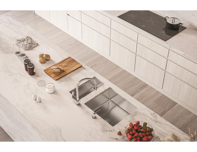 Svetla moderna kuhinja z velikim otokom in marmornatim pultom, v katerega je vgrajeno inox pomivalno korito z dvema posodama in kromirano armaturo; na pultu so lesena deska z rogljički, kozarci, jajca in skleda s svežimi jagodami.
