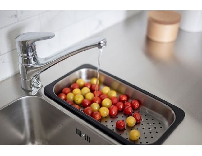 Detajl kuhinjskega pomivalnika z inox koritom in kromirano armaturo, iz katere teče voda v perforirano posodo za odcejanje; v njej so oprani rdeči in rumeni češnjevi paradižniki.
