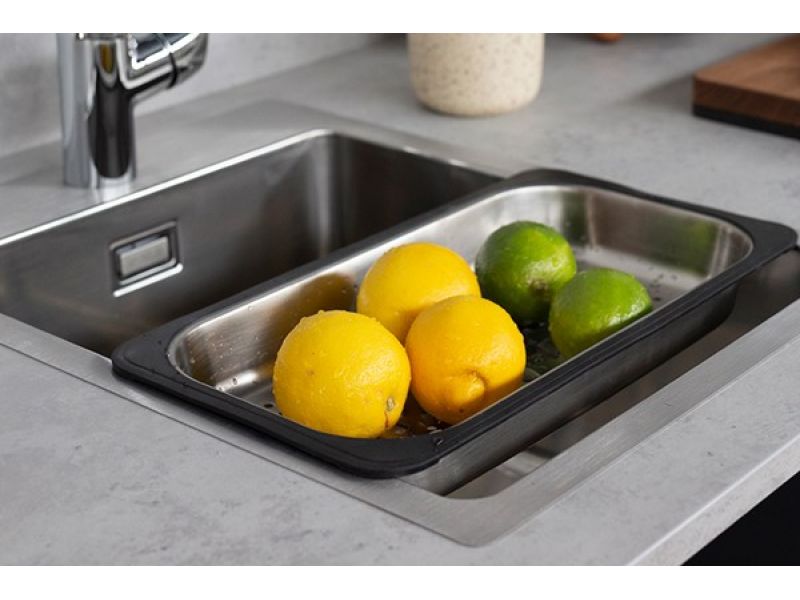 Inox kuhinjsko korito z vstavljeno odcejalno posodo s silikonsko obrobo, v katerem so limone in limete; ob koritu je kromirana armatura, vse skupaj pa je vgrajeno v svetel kuhinjski pult.