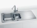 Inox kuhinjsko pomivalno korito z dvema posodama različnih velikosti in integrirano odcejalno površino ter kromirano armaturo, vgrajeno v bel kuhinjski pult.