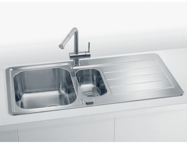 Inox kuhinjsko pomivalno korito z dvema posodama različnih velikosti in integrirano odcejalno površino ter kromirano armaturo, vgrajeno v bel kuhinjski pult.