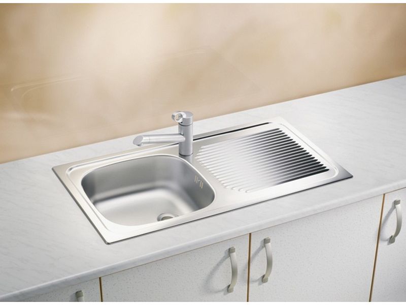 Inox kuhinjsko pomivalno korito z eno posodo in rebrasto odcejalno površino ter kromirano enoročno armaturo, vgrajeno v svetel kuhinjski pult nad belimi omaricami.
