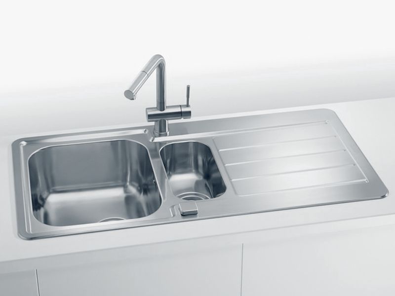 Inox kuhinjsko pomivalno korito z dvema posodama različnih velikosti in integrirano odcejalno površino ter kromirano armaturo, vgrajeno v bel kuhinjski pult.